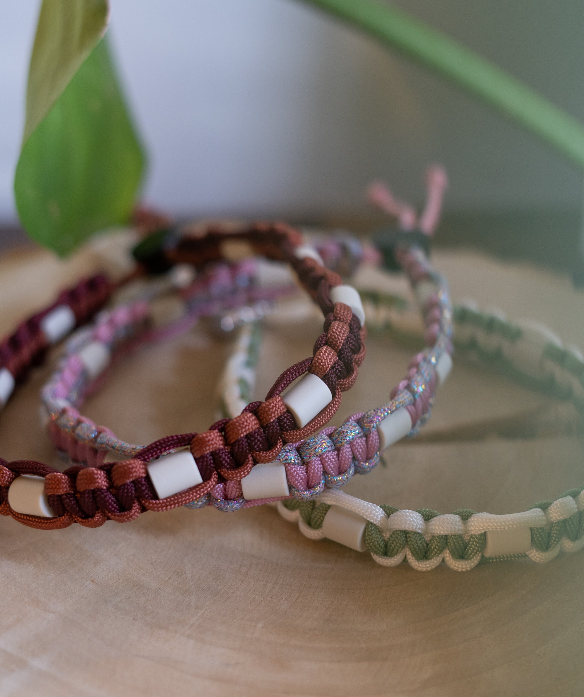 Assortiment de trois colliers anti-tiques naturels pour chien posés sur une tranche de bois. On distingue de haut en bas : un modèle bordeaux (rouille), un modèle rose avec fil pailleté brillant, et un modèle vert sauge. Tous intègrent des perles en céramique EM dans leur tressage artisanal.