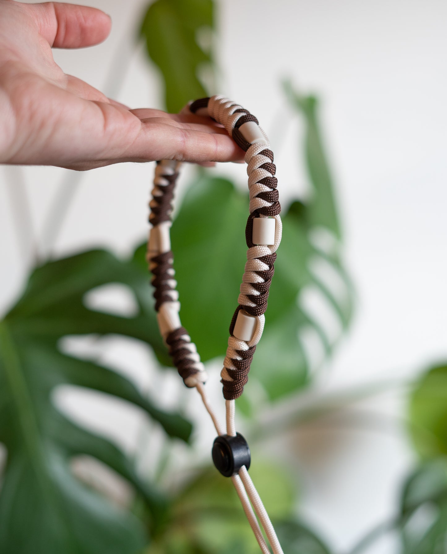 Gros plan sur un collier anti-tiques naturel pour chien tenu à la main. Le collier est tressé en paracorde marron et beige et intègre des perles en céramique EM pour repousser les parasites. Arrière-plan flou avec des feuilles de Monstera soulignant le côté écologique et sans produits chimiques du collier.